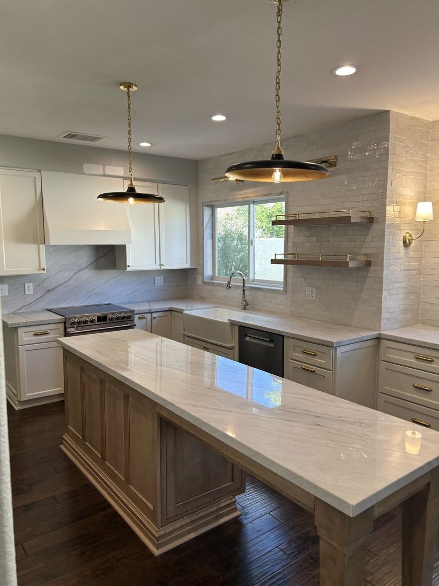 A modern kitchen featuring white cabinets, a large marble countertop island, stainless steel appliances, and pendant lighting. The decor includes light-colored tiles on the walls and wooden flooring.